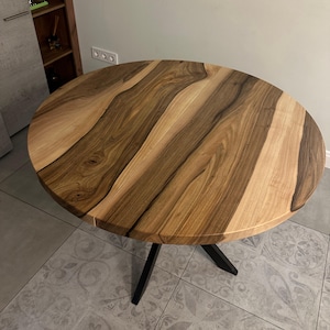 May include: A round dining table featuring a natural wood grain tabletop with alternating light and dark brown tones. The table is supported by a black metal base with three angled legs that meet at a central point beneath the circular surface.