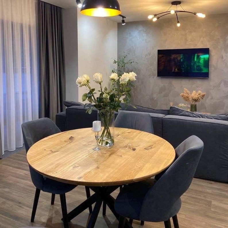 May include: A round light-colored wooden dining table with a black metal base, surrounded by four dark gray upholstered chairs. A clear glass vase filled with white roses and a single white pillar candle in a glass holder sit on the table.