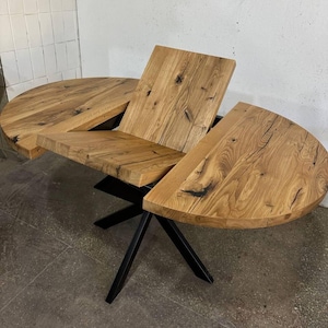 Op de afbeelding: Ronde eettafel van lichtbruin hout met een natuurlijke afwerking. De tafel is uitklapbaar. De tafel heeft een zwart metalen onderstel. Het tafelblad heeft zichtbare houtnerf en donkere knopen. Uitklapbaar.