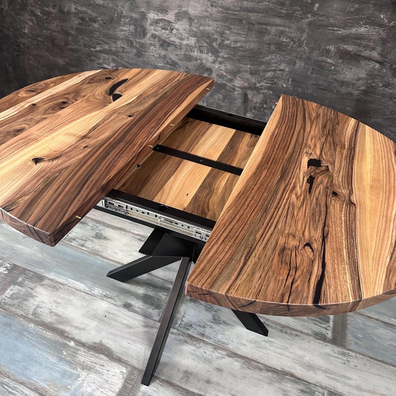 May include: A round wooden dining table with a natural grain finish and dark knots, featuring a split top design that reveals an internal extension leaf. The table is supported by a black metal base with angled legs and includes a sliding mechanism.