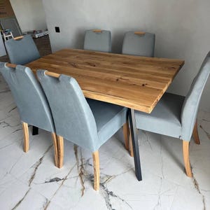 Op de afbeelding: Een rechthoekige houten eettafel met een natuurlijke houten afwerking, gecombineerd met zes grijs gestoffeerde stoelen. De stoelen hebben lichte houten poten en armleuningen. De tafel heeft zwarte metalen poten. De stoelen zijn rond de tafel geplaatst.