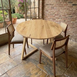 May include: A round light-colored wooden dining table with a geometric base, accompanied by three chairs featuring dark wood frames, woven cane backrests, and light gray upholstered seats, arranged on a stone floor.