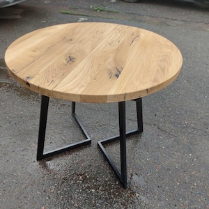 May include: A round table featuring a light brown natural wood grain top with visible knots and dark fissures. The table is supported by a geometric base constructed from four black metal legs that form two interconnected angular frames.