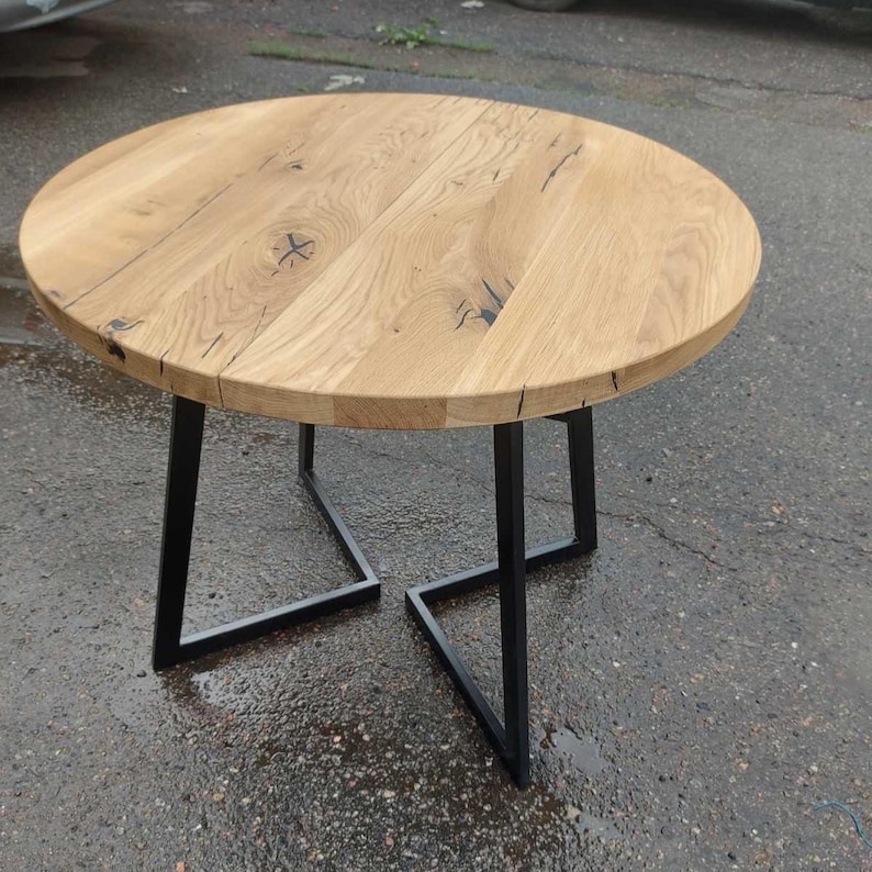 May include: A round table featuring a light brown natural wood grain top with visible knots and dark fissures. The table is supported by a geometric base constructed from four black metal legs that form two interconnected angular frames.