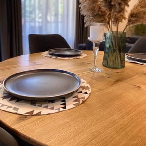 May include: A light oak wood dining table set with two matte black round dinner plates on patterned placemats. A clear glass vase with tall dried pampas grass and a white pillar candle in a glass pedestal holder sit in the center of the table.
