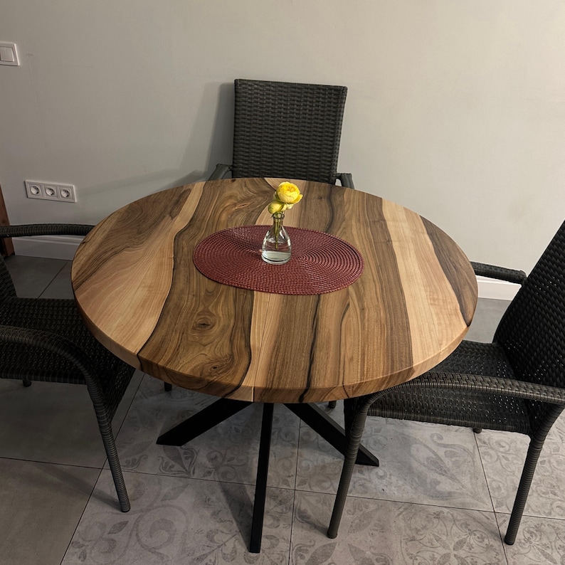May include: A round wooden dining table with a natural live edge grain pattern and a black metal base, accompanied by three dark gray woven chairs. A small clear glass vase with yellow flowers sits on a round red textured placemat on the table.