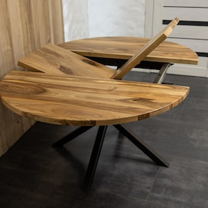 Puede incluir: Una mesa de comedor redonda de madera con un acabado de veta de madera marrón claro natural. La mesa cuenta con un mecanismo de extensión central actualmente en posición entreabierta, apoyada sobre una base de metal negro con cuatro patas.