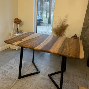 K&ouml;nnte beinhalten: Ein rechteckiger Holztisch mit einem gestreiften Muster in Braun- und Beiget&ouml;nen. Der Tisch hat schwarze Metallbeine. Der Tisch ist etwa 152 cm lang und 76 cm breit.