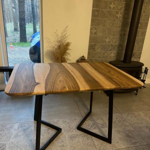 K&ouml;nnte beinhalten: Ein rechteckiger Holztisch mit nat&uuml;rlicher Holzmaserung. Die Tischplatte wird von schwarzen Metallbeinen getragen. Der Tisch ist etwa 122 cm lang und 91 cm breit.