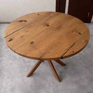 Op de afbeelding: Een ronde houten tafel met een natuurlijke afwerking. Het tafelblad is gemaakt van planken met zichtbare houtnerf en donkere accenten. De tafel heeft een driepoot. De tafel heeft een diameter van ongeveer 100 cm.