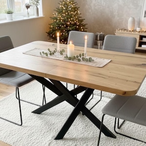May include: A rectangular light wood dining table with a black metal X-shaped base. The table is surrounded by four gray upholstered chairs with thin black metal legs. A beige table runner holds two tall white candles and a green leaf garland.