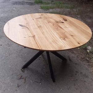 May include: A round dining table featuring a light-colored natural wood grain top with visible knots and organic patterns. The tabletop is supported by a black metal base consisting of four angled legs that meet in a central point beneath the surface.