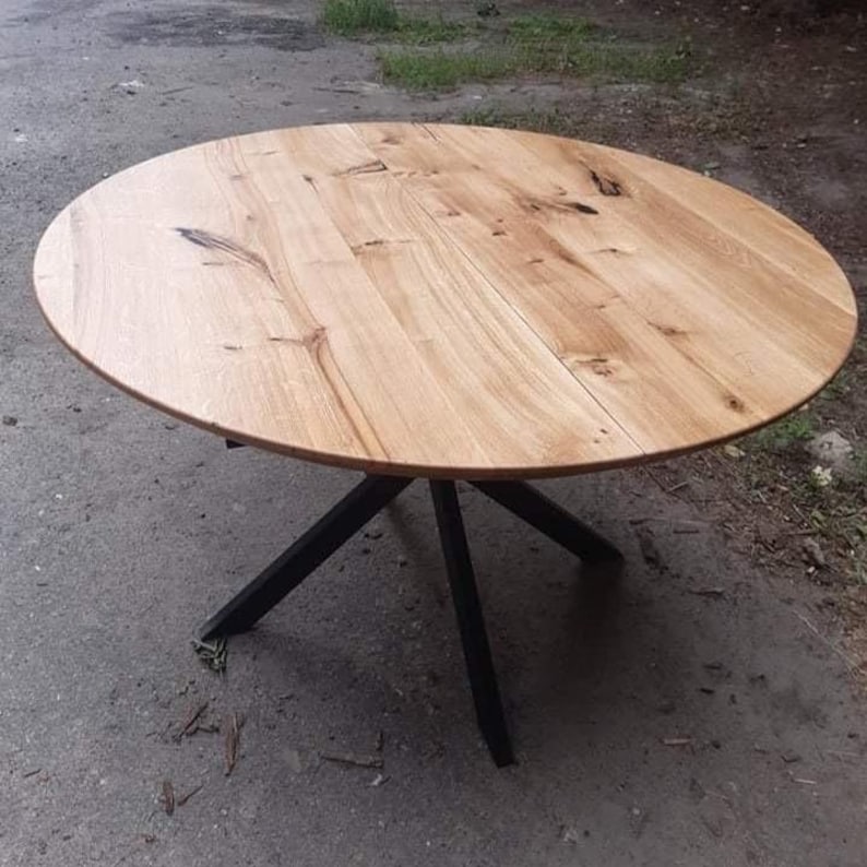 May include: A round dining table featuring a light-colored natural wood grain top with visible knots and organic patterns. The tabletop is supported by a black metal base consisting of four angled legs that meet in a central point beneath the surface.