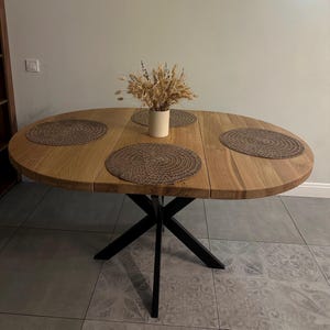 K&ouml;nnte beinhalten: Ein ovaler Holzesstisch mit nat&uuml;rlicher hellbrauner Maserung, der auf einem schwarzen Metallkreuzgestell steht. Die Tischplatte ist mit drei runden, geflochtenen braunen Platzsets und einer cremefarbenen Vase mit getrockneten Weizen&auml;hren dekoriert.