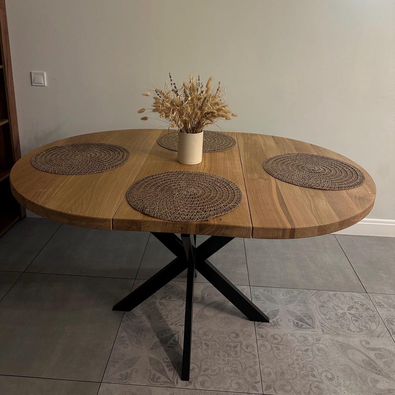 K&ouml;nnte beinhalten: Ein ovaler Holzesstisch mit nat&uuml;rlicher hellbrauner Maserung, der auf einem schwarzen Metallkreuzgestell steht. Die Tischplatte ist mit drei runden, geflochtenen braunen Platzsets und einer cremefarbenen Vase mit getrockneten Weizen&auml;hren dekoriert.