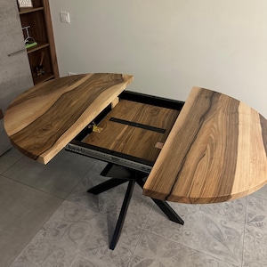 May include: A round dining table with a natural wood grain top featuring a split design that reveals an internal extension mechanism. The table has a black metal base with a central support structure and four angled legs extending outward.