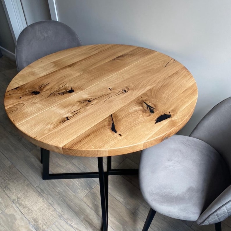 May include: A round wooden dining table with a natural light brown grain and visible dark knots, supported by a black metal cross-base frame. Two grey upholstered chairs with curved backrests are positioned on opposite sides of the table.