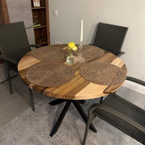 May include: A round dining table with a natural wood grain top featuring light and dark brown tones, supported by a black metal base. Four dark gray woven chairs surround the table, which is set with four round brown placemats, a glass vase with yellow flowers, and a tall white taper candle in a clear glass holder.
