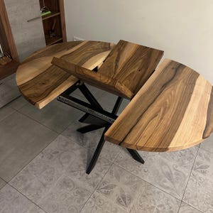May include: A round dining table with a natural wood grain top featuring a live edge and a central extension mechanism. The table is supported by a black metal base with angled legs and includes a sliding track system for the expandable leaf section.