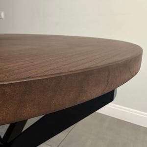 Peut inclure: Gros plan sur une table ronde en bois avec une finition marron fonc&eacute;. La table a un dessus &eacute;pais et solide et une base en m&eacute;tal noir. Le grain du bois est visible sur la surface.