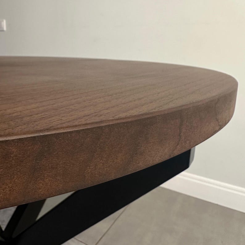 Peut inclure: Gros plan sur une table ronde en bois avec une finition marron fonc&eacute;. La table a un dessus &eacute;pais et solide et une base en m&eacute;tal noir. Le grain du bois est visible sur la surface.