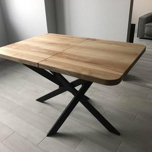 Pode incluir: Uma mesa de jantar retangular com um tampo de madeira natural de tom claro, cantos arredondados e uma costura central visível. A mesa é suportada por uma base de metal preto composta por pernas diagonais cruzadas em forma de X.
