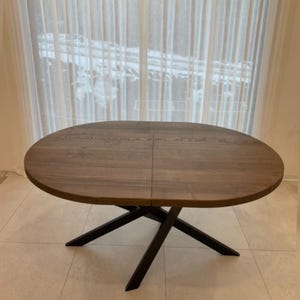 Pode incluir: Uma mesa de jantar redonda de madeira com um padrão visível de veios de madeira castanho-escuro na superfície. A mesa apresenta uma costura central que indica um design extensível e é suportada por uma base de metal preto com quatro pernas.