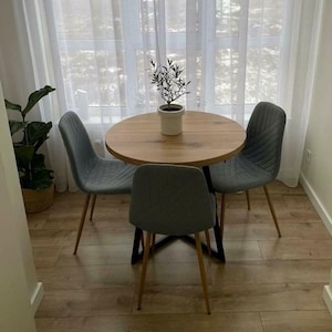 May include: A round wooden dining table with a light brown finish and a black metal base, accompanied by three grey upholstered chairs with vertical stitching and thin metal legs. A small potted olive tree sits in the center of the table.