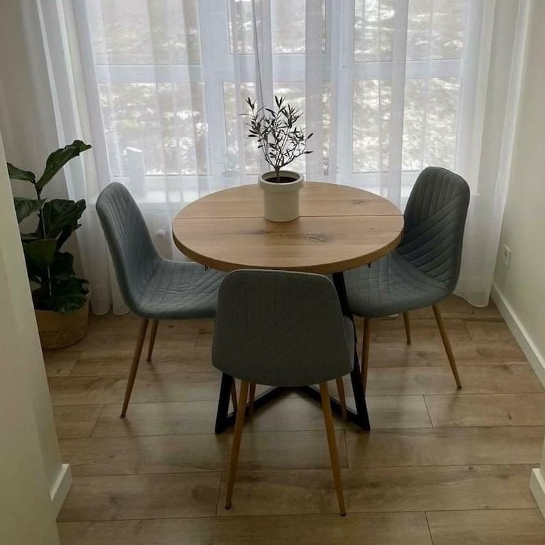 May include: A round wooden dining table with a light brown finish and a black metal base, accompanied by three grey upholstered chairs with vertical stitching and thin metal legs. A small potted olive tree sits in the center of the table.