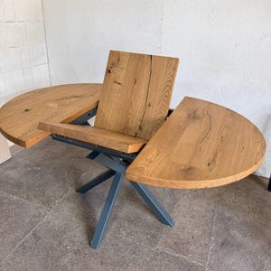 Op de afbeelding: Een uitschuifbare eettafel van lichtbruin hout met een natuurlijke afwerking. De tafel heeft een donkergrijs metalen onderstel en een centrale plaat. Het tafelblad toont zichtbare houtnerf en een uniek ontwerp. De tafel is ontworpen voor meerdere personen.