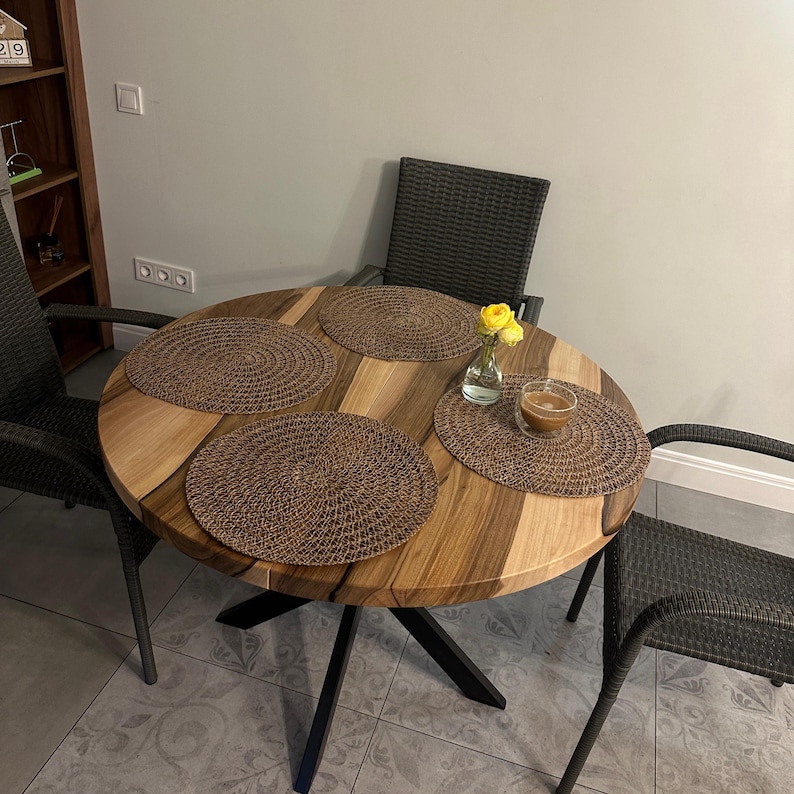 May include: A round wooden dining table with a natural grain pattern and black metal legs, accompanied by three dark gray woven chairs. The table is set with four circular brown woven placemats, a small glass vase with yellow flowers, and a glass cup.