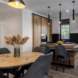May include: A dining area featuring a round wooden table with a glass vase of pampas grass, surrounded by four dark gray upholstered chairs. In the background, a kitchen with wood-grain cabinetry, black countertops, and three black pendant lights.