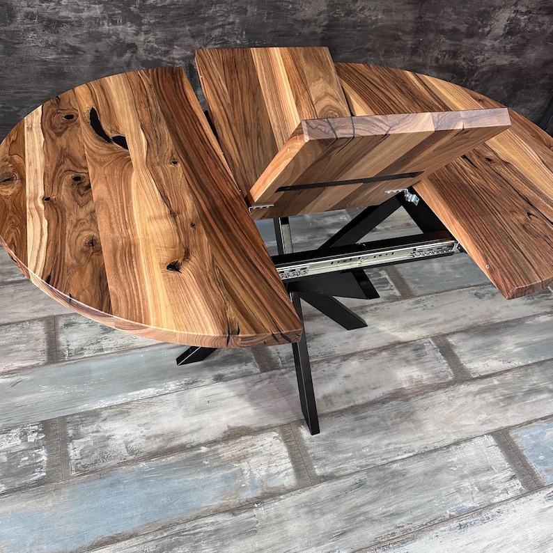 May include: A round wooden dining table with a natural grain pattern and dark knots, featuring an extendable center leaf mechanism. The table is supported by a black metal base with angled legs and visible silver metal sliding tracks.