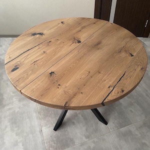 May include: A round dining table featuring a natural light brown wood grain surface with visible dark knots and cracks filled with black resin. The tabletop is supported by a black metal base with angled legs, providing a stable foundation.