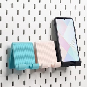 Op de afbeelding: Drie kleine, geribbelde telefoonstandaards in teal, lichtroze en zwart, gemonteerd op een wit geperforeerd bord. De zwarte standaard houdt een smartphone met een lichtgekleurd scherm vast. Elke standaard heeft een U-vormige haak voor ondersteuning.