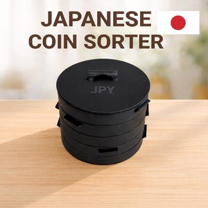 May include: A black cylindrical Japanese coin sorter with a stackable design. The top lid features a small handle and the text JPY embossed in the centre. The unit consists of multiple circular tiers with small side slots for organising currency.