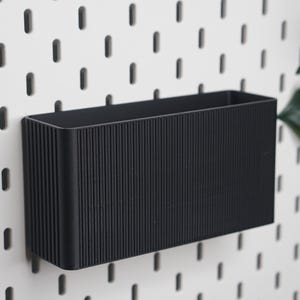 May include: A rectangular black storage bin with a vertical ribbed texture on the exterior. The container features an open top and is designed to be mounted onto a white pegboard panel with a repeating pattern of vertical slots for organisation.