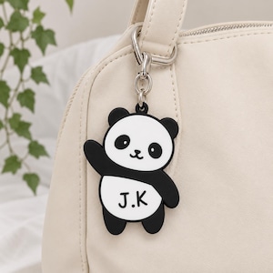 Puede incluir: Un llavero con forma de panda en blanco y negro que presenta las letras J.K impresas en su vientre. El dije está sujeto a un mosquetón y una argolla de metal plateado, enganchados al asa de un bolso de mano de color crema.