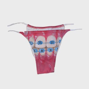 May include: A novelty thong featuring a printed image of a mouth with teeth and blue braces. The thong has white string straps and a red and white waistband. The graphic design is a close-up of a mouth with healthy teeth and braces.