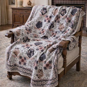 Könnte beinhalten: Eine gesteppte Tagesdecke mit einem Patchwork-Design aus Blumen- und Karomustern in den Farben Altrosa, Anthrazit und Creme. Der Stoff weist eine sichtbare Steppstruktur sowie eine dekorative Bordüre entlang der Ränder der Decke auf.