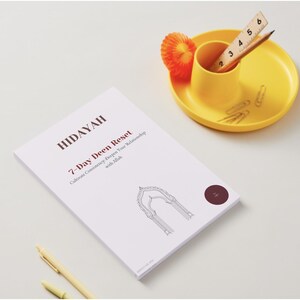 May include: A white book titled "HIDAYAH 7-Day Deen Reset" with an archway graphic. A yellow desk tidy contains a ruler, pencil, and paperclips. An orange decorative item is also present on a white surface.