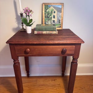 May include: A small, antique wooden side table with a single drawer and two round knobs. The table is stained a rich reddish-brown color. A framed painting, books, a candle, and a potted orchid are displayed on top. The table's legs are turned and tapered.