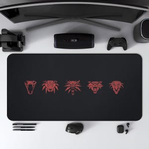 May include: Black rectangular desk mat featuring five red stylized animal emblems. A computer mouse, pens, and a speaker are also visible. A curved monitor, VR headset, and game controller are also present.