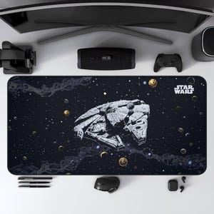 May include: A black rectangular desk mat featuring a detailed illustration of the Millennium Falcon spaceship against a starry space background. The Star Wars logo is in the upper right corner. Other items include a computer monitor, speakers, and a mouse.