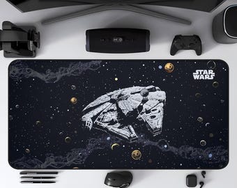 May include: A black rectangular desk mat featuring a detailed illustration of the Millennium Falcon spaceship against a starry space background. The Star Wars logo is in the upper right corner. Other items include a computer monitor, speakers, and a mouse.
