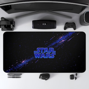 May include: A black rectangular desk mat featuring the Star Wars logo in blue. The mat has a galaxy-themed design with blue and purple accents. Other items on the white surface include a computer monitor, speakers, and a mouse.