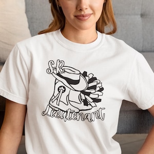 Puede incluir: Camiseta blanca con un gráfico negro de un sombrero de vaquero, botas y un pompón, con la palabra "Lieutenant" en cursiva. La camiseta es de cuello redondo y la lleva una persona.