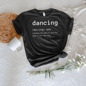 Könnte beinhalten: Schwarzes T-Shirt mit weißem Text, der lautet: "dancing (dan-cing) Verb Ermöglicht es der Musik, den Körper zu bewegen, um die Geschichte zu erzählen."