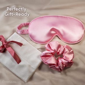 May include: A pink satin sleep mask, scrunchie, and a white pouch with a pink ribbon. The sleep mask and scrunchie are a matching light pink color. The text "Perfectly Gift-Ready" is in the upper left corner.