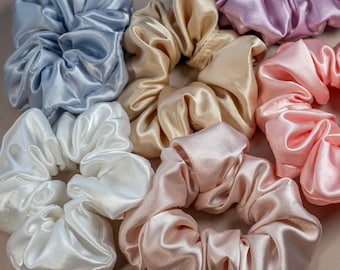 May include: A collection of six satin hair scrunchies in light blue, champagne, white, lavender, pink, and dusty rose. Each scrunchie features a gathered, pleated fabric design with a smooth, reflective finish. The brand name Ammaliante is printed in the bottom left corner.